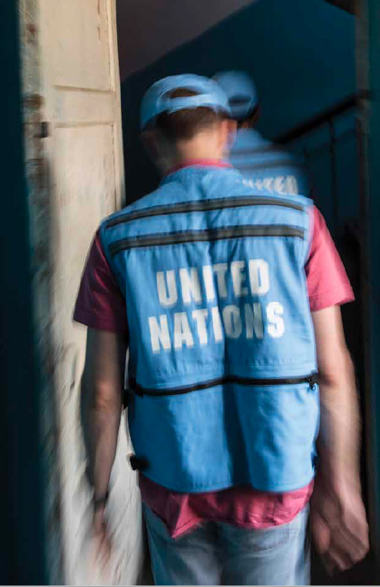 Blurred image showing two United Nations staff members walking into a run-down building with scuffed blue walls and visible posted notices.