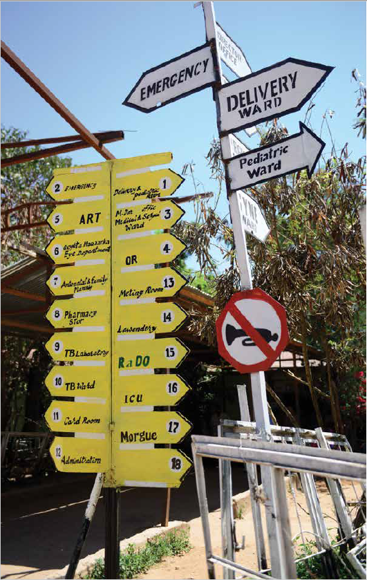 Outdoor hospital signage featuring multiple yellow and white arrow signs directing visitors to various medical units such as the ICU, Pharmacy, TB Laboratory, and ART department. A red and white no-horn sign is in the foreground, with trees and metal railings nearby.