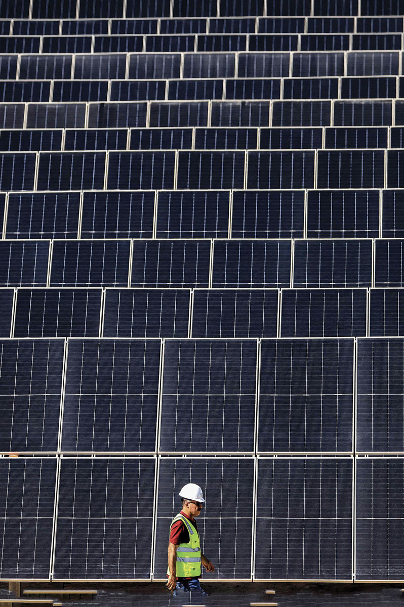 A worker in a reflective vest and hard hat walks in front of a large wall covered with rows of solar panels.