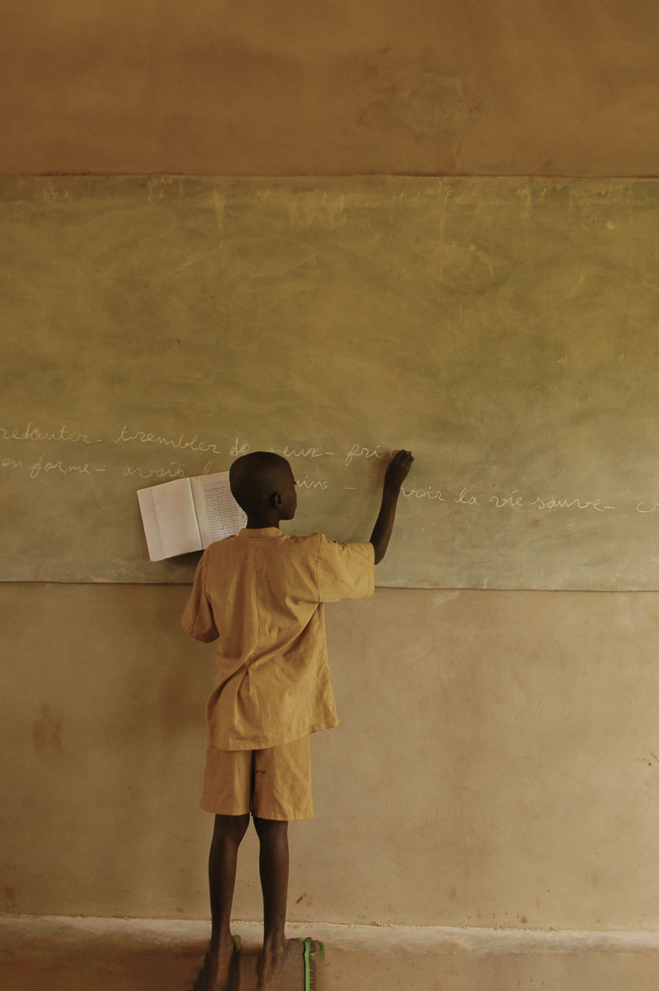 Boy dressed in yellow writes French words on a chalkboard, propping up a workbook as he stands on a ruler to reach the writing surface.
