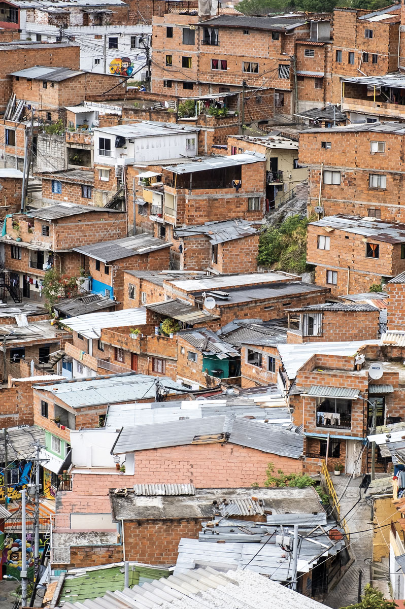 An urban settlement built on a slope, featuring clusters of rustic, unfinished red brick houses with corrugated metal rooftops and scattered bright patches of painted walls.