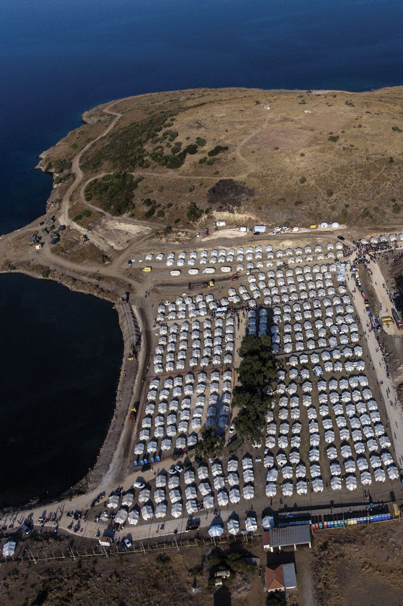 Aerial view of a coastal refugee camp, showing hundreds of white tents arranged in neat rows on dry, brown land beside the deep blue sea, with some permanent structures and roads interspersed.