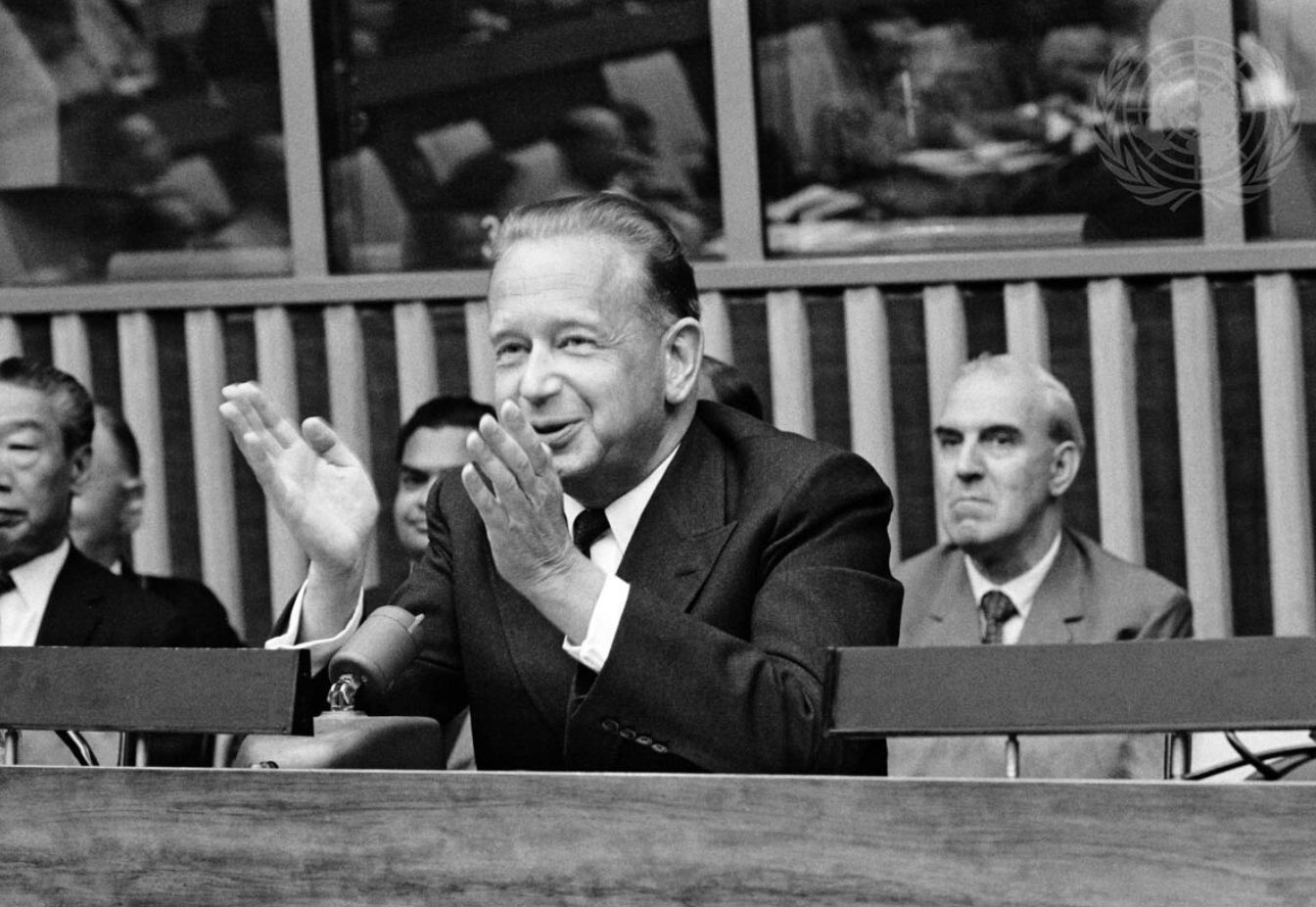Black-and-white photo of Dag Hammarskjöld, the former Secretary-General, gesturing with open palms while addressing a formal gathering, with several other men seated behind him.