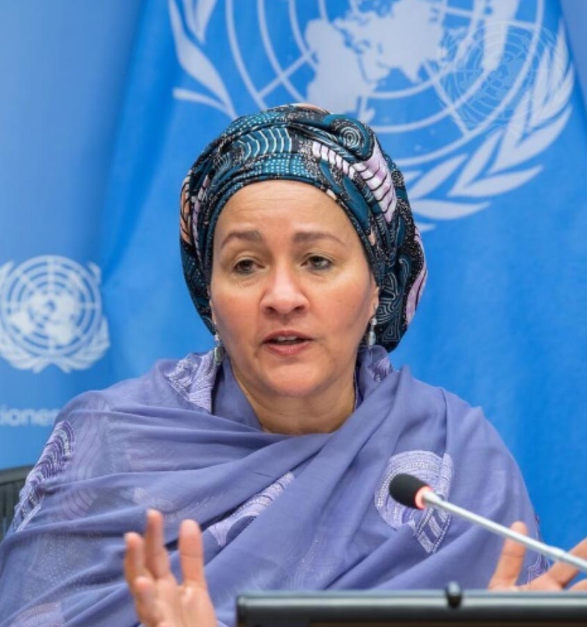 Ms. Amina Mohammed, Deputy Secretray-General, in a purple and blue traditional headscarf and shawl speaks at a United Nations press conference, seated in front of a blue UN flag with a microphone in front of her.