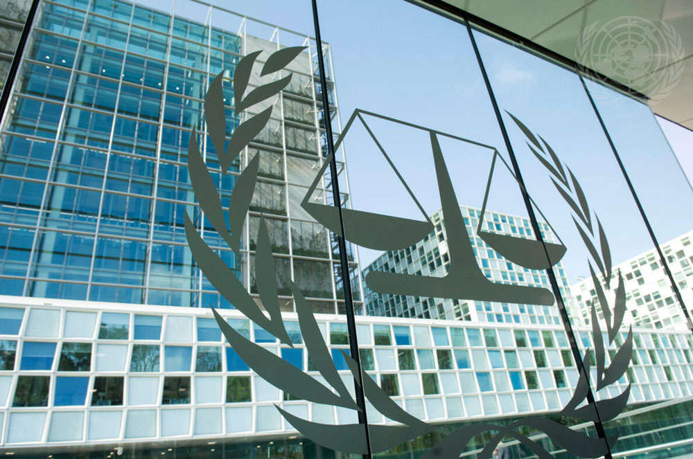 A glass facade of a modern building reflects the emblem of the International Criminal Court, featuring a pair of balanced scales surrounded by laurel branches.