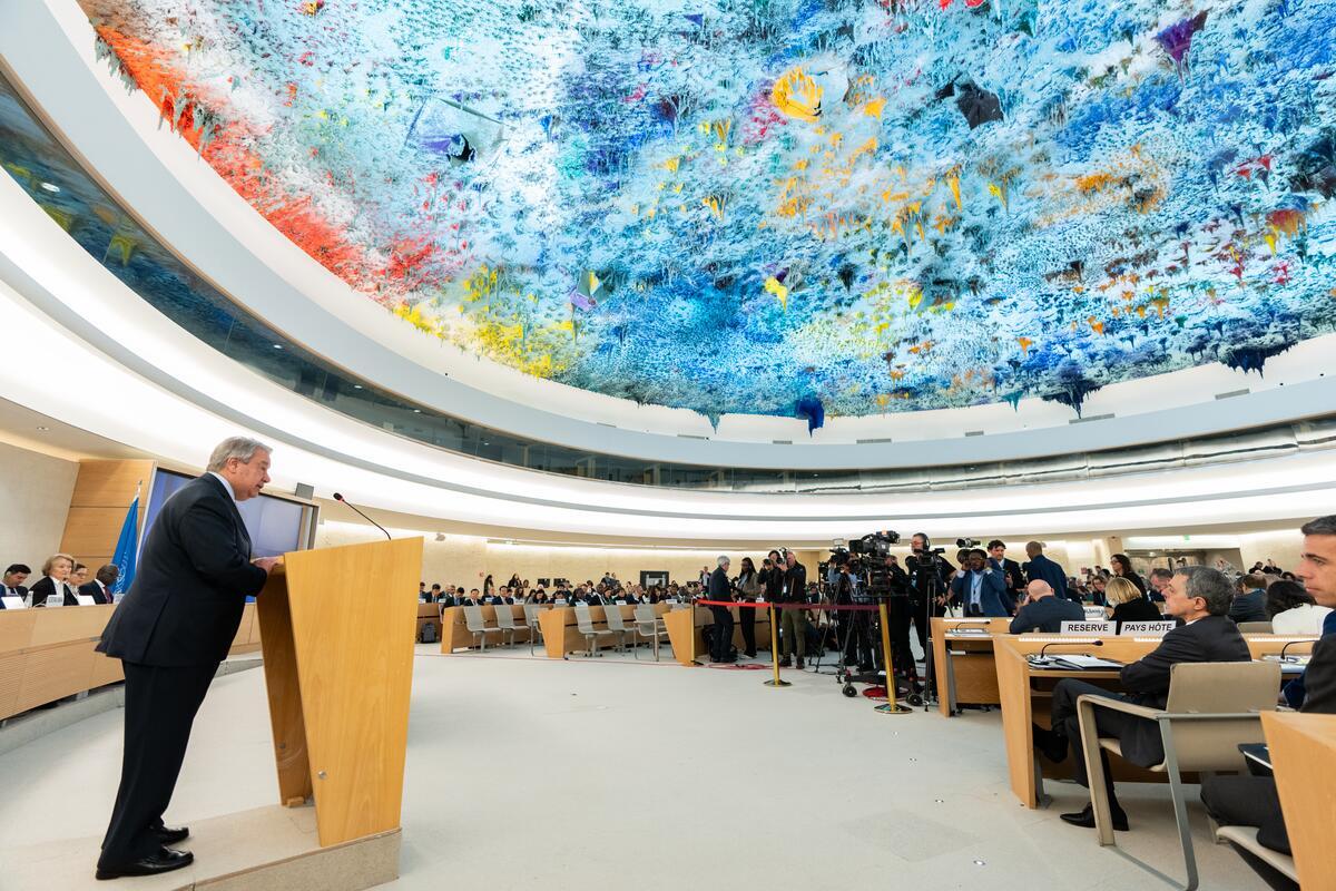 A wide view of a diplomatic assembly shows Secretary-General António Guterres delivering a speech at a wooden podium, with international delegates seated and cameras set up beneath the stunning blue and multicolored ceiling artwork of the Human Rights Council conference hall.