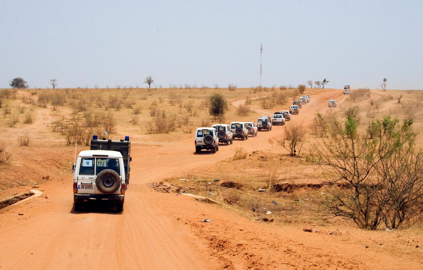 A convoy of white off-road vehicles, some marked with "UN," moves across an arid, open terrain dotted with small shrubs and isolated trees.