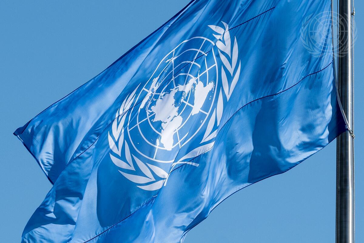 A bright blue flag featuring the emblem of the United Nations—a world map surrounded by olive branches—waving against a clear blue sky.